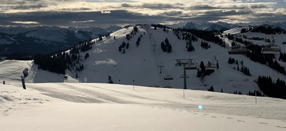 Snedækkede skiløjper i Westendorf under en delvist overskyet himmel, med skilifter langs bakken. Solen kigger gennem skyerne og kaster et blødt lys på det bjergrige landskab i baggrunden – en perfekt ramme om en mindeværdig skiferie.