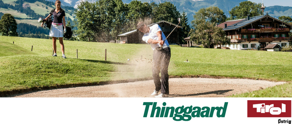 En golfspiller svinger sig i en sandfælde på en frodig, grøn bane med bjerge i baggrunden, der minder om den naturskønne skønhed, der findes nær Wagrain. En kvinde står i nærheden og ser på. Logoer for Thinggaard og Tirol er vist nederst på billedet.