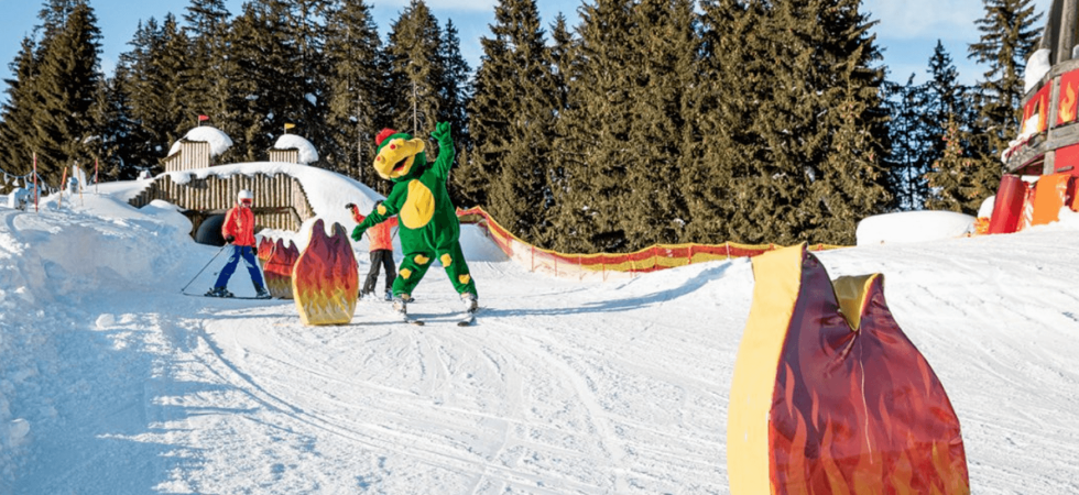 En person i et grønt dinosaurkostume står på ski ned ad en sneklædt skråning på deres skiferie, omgivet af flammeformede dekorationer. To andre skiløbere følger efter, silhuet mod de høje fyrretræer og den klare blå himmel i Wagrain.