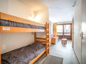 Et lille, minimalistisk værelse perfekt til et skiferie-retreat, med trækøjesenge til venstre og en lyserød sofa ved de store vinduer til højre. En trækiste fungerer som sofabord. Det lyse trægulv og de neutrale vægge soler sig i blød belysning og byder på fredfyldte vibes, der minder om Saalbach og Westendorfs hyggelige charme.