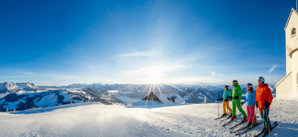En gruppe skiløbere i livlige outfits står på en sneklædt bjergskråning og nyder deres skiferie i Westendorf under en klar blå himmel og strålende solskin. En lille bygning og den vidtstrakte bjergkæde skaber en perfekt kulisse.