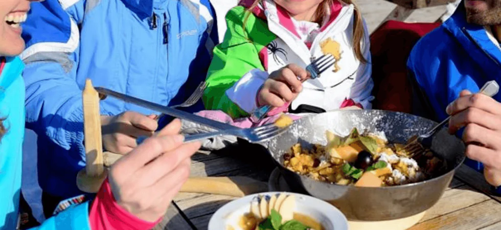 En familie klædt i farverige vinterjakker nyder et måltid udendørs under deres skiferie. De sidder ved et træbord med en pande med kogt mad og en skål suppe. Sne kan ses i baggrunden, og alle smiler og holder redskaber og nyder denne hyggelige stund sammen.