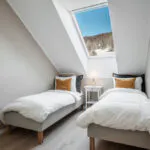Et hyggeligt loftsværelse i Saalbach har to enkeltsenge med hvidt sengetøj og gule accentpuder. Et lille hvidt natbord mellem sengene rummer en lampe, mens skråloftets ovenlys giver en betagende udsigt over et snelandskab og blå himmel.
