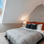 Hyggeligt loftssoveværelse med loft til kip og ovenlys, der minder om et Wagrain skiferie retreat. Sengen har en tuftet sengegavl, gråt sengetøj og orange og mørkegrå puder. To hvide natborde med lamper tilføjer charme, mens vinduet afslører et snelandskab udenfor.