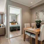 Et hyggeligt opholds- og spiseområde med beige vægge, med et træbord med hvide stole og en vase med blomster. Gennem en åben dør forstærker et glimt af et soveværelse med et grønt sengetæppe skiferiestemningen. Store vinduer viser Wagrains sneklædte bjergudsigt.
