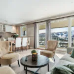 En lys stue med beige stole, et hvidt spisebord og en sofa. Store vinduer afslører en sneklædt bjergudsigt over Saalbach. Indretningen er neutral med grønne og træaccenter, hvilket skaber en hyggelig atmosfære perfekt til planlægning af dit næste skiferieeventyr.