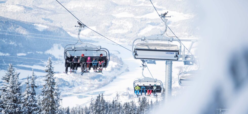 Et skisportssted udfolder sig, mens folk i stolelifte stiger op over Wagrains sneklædte landskab. Træer er støvet med sne, og fjerne bjerge danner baggrunden. Den klare himmel forstærker den vinterlige, fredelige atmosfære, og inviterer skirejseentusiaster til at fordybe sig i dette alpine paradis.