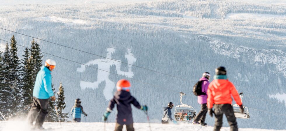 Skiløbere i alle aldre navigerer på en sneklædt skråning i Wagrain med en naturskøn kulisse af snedækkede bakker og fyrretræer. En skilift er synlig i det fjerne under en klar himmel. Nogle personer bærer farverige jakker og hjelme.