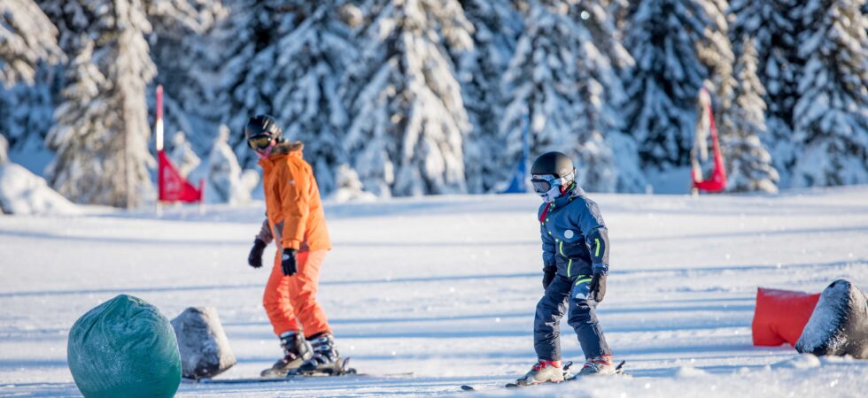 To personer står på ski på en sneklædt skråning i Saalbach, omgivet af snedækkede træer. Den ene bærer et orange outfit og den anden et mørkt. Begge har hjelme og beskyttelsesbriller, med farverige flag i baggrunden, der fanger den spændende essens af skirejser.