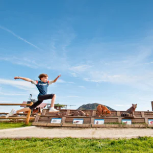 Et barn i blå skjorte og shorts hopper energisk over et plankeværk i et åbent græsareal under deres Sommerferie i Westendorf. I baggrunden er træudskæringer af forskellige dyr sat på baggrund af et bjerg under en klar blå himmel.
