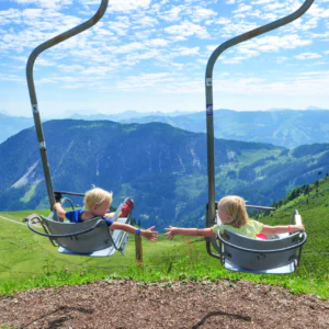 To børn i stoleliften rækker ud mod hinanden, mod det naturskønne bjerglandskab i Westendorf. Den delvist overskyede himmel og de pulserende grønne græsklædte bakker nedenfor fanger essensen af et perfekt sommerferieeventyr.