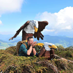 En person i en plaidskjorte sidder i en stor rede på et bjerg, omfavnet af to ørneskulpturer i træ. Det er de perfekte rammer for en "Sommerferie i Westendorf", med klar blå himmel og grønne bakker, der maler en idyllisk kulisse.