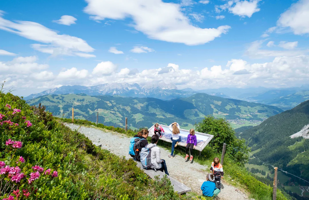 En naturskøn udsigt over bjergene under en Sommerferie i Westendorf, hvor en gruppe vandrere holder en pause ved et stiskilt. Pink blomster og grønt omkranser stien under blå himmel med spredte skyer, mens vidtstrakte dale og bjerge strækker sig i det fjerne.