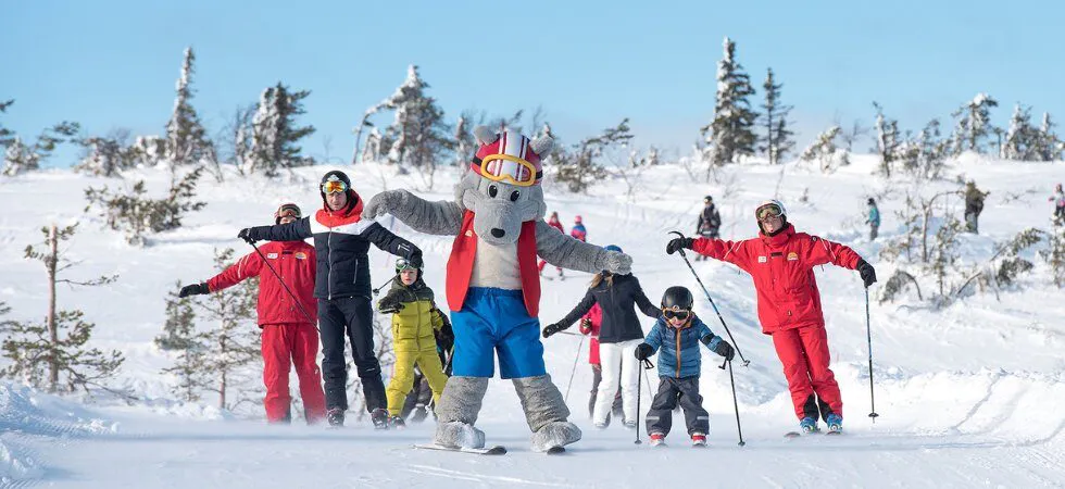 En gruppe mennesker på ski ned ad en sneklædt skråning under deres skiferie med en stor grå tegneseriefigurmaskot i farverigt skiudstyr, der fører dem i maleriske Westendorf. Baggrunden har snedækkede træer og en klar blå himmel.