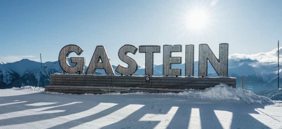 Et stort skilt, der staver GASTEIN står på en snedækket platform under en klar himmel. Det er en indbydende scene for skirejseentusiaster, mens solen skinner klart over fjerne bjerge og kaster lange skygger på den snedækkede jord, der minder om Saalbachs fortryllende vinterlandskaber.