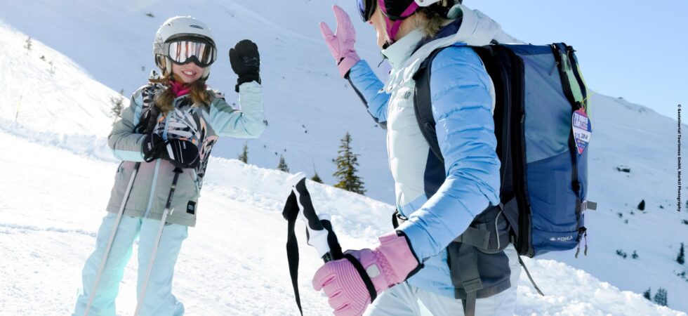 To skiløbere, iført hjelme og briller, deler en high-five på en sneklædt skråning under deres skiferie. Den ene er en voksen med rygsæk og skistave, den anden et barn. De griner midt i Westendorfs solbeskinnede bjerge under en klar blå himmel.