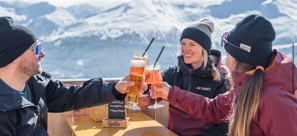 Tre personer i vintertøj klirrer i glas ved et udendørs bord mod de sneklædte bjerge i Wagrain. De smiler og nyder drinks med skarpt sollys, der fremhæver scenen, og fanger et perfekt øjeblik af glæde midt i Østrigs fantastiske vinterlandskab.