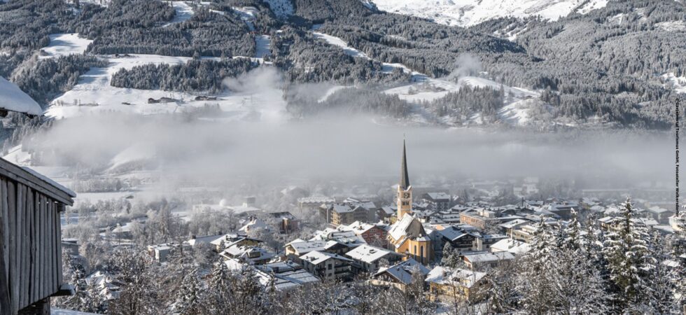 Et vinterlandskab af en snedækket landsby med et fremtrædende kirketårn. Bjerge og skove, perfekt til skiferie, er dækket af sne under en klar blå himmel. Et lag lavthængende skyer lægger sig over landsbyen og skaber en fredfyldt atmosfære ideel for skirejser.