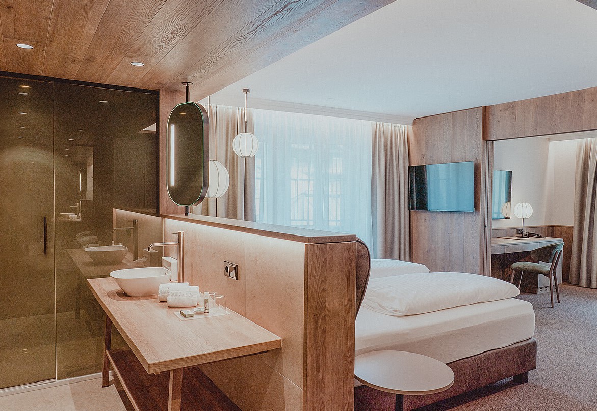 Et moderne hotelværelse med trædesign med en dobbeltseng, et vægmonteret tv, et skrivebord med en stol og et synligt åbent badeværelsesområde med en glasparti. Varm belysning og store vinduer med gardiner bidrager til den hyggelige stemning.