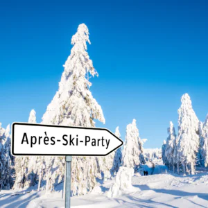 Et snelandskab med høje snedækkede træer under en klar blå himmel. I forgrunden står der på et skilt Après-Ski-Party, der peger mod det fjerne, og inviterer dig til en afterski social sammenkomst fyldt med varme og kammeratskab ud over pisterne.
