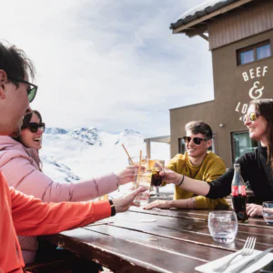 En gruppe på fire personer, pakket ind i varmt tøj, sidder ved et udendørs bord og soler sig i det solrige afterski-skær. De klirrer glade glas, med sneklædte bjerge, der giver en fantastisk baggrund for deres latter og kammeratskab.