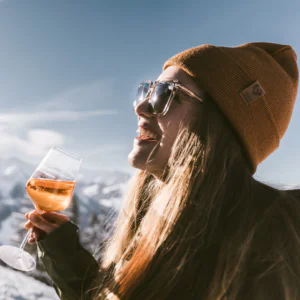 En person i en brun hue og solbriller smiler, mens han holder et glas vin udendørs. Baggrunden byder på snedækkede bjerge og en klar blå himmel, der fanger den livlige afterski-atmosfære.