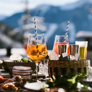 Et bord dækket til udendørs nydelse med to cocktails garneret med stribede sugerør, en kurv med metalkopper og snacks inkarnerer perfekt afterski-stemningen. Sneklædte bjerge og en klar himmel danner en fantastisk kulisse, der skaber en hyggelig, festlig atmosfære.