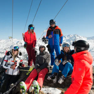 En gruppe på seks skiløbere i farverigt udstyr sidder og står på en sneklædt bjergskråning under en klar blå himmel. For at fejre Nytår på ski, er de i nærheden af en skilift, nyder en pause med drinks, omgivet af naturskønne snedækkede tinder.
