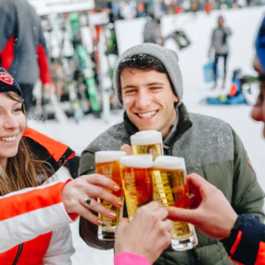 I sneklædte udendørs omgivelser hæver en gruppe mennesker i vintertøj deres ølglas for at skåle. De smiler, omgivet af ski, jakker, hatte og handsker. Det er virkelig Nytår på ski, der fanger den festlige og muntre stemning perfekt.