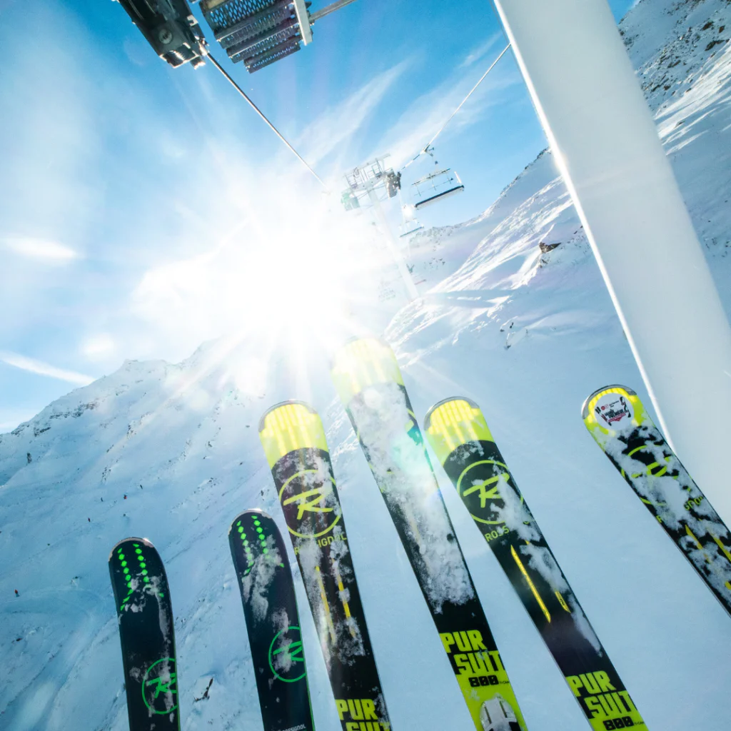 Nærbillede af flere par ski, der peger opad under en solrig, klar blå himmel. En skilift er synlig ovenfor, med snedækkede bjerge i baggrunden. Sollyset skaber et skarpt skær, der understreger det vinterlige landskab – perfekt til at fejre Nytår på ski.