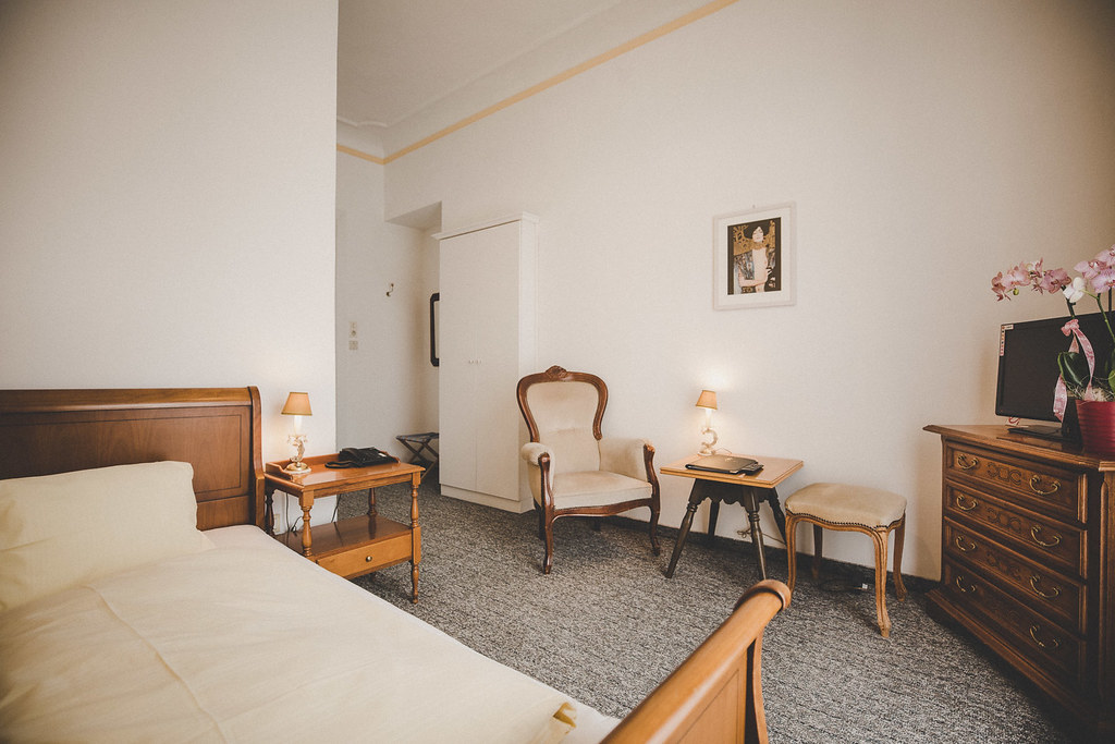 Et hyggeligt hotelværelse med en enkelt træseng med et gult sengetæppe, en vintage lænestol og en kommode i træ. Et lille bord med en lampe og en telefon sidder ved siden af sengen. Et tv er på kommoden, og en lyserød orkidé er i nærheden af det. Et maleri hænger på væggen.
