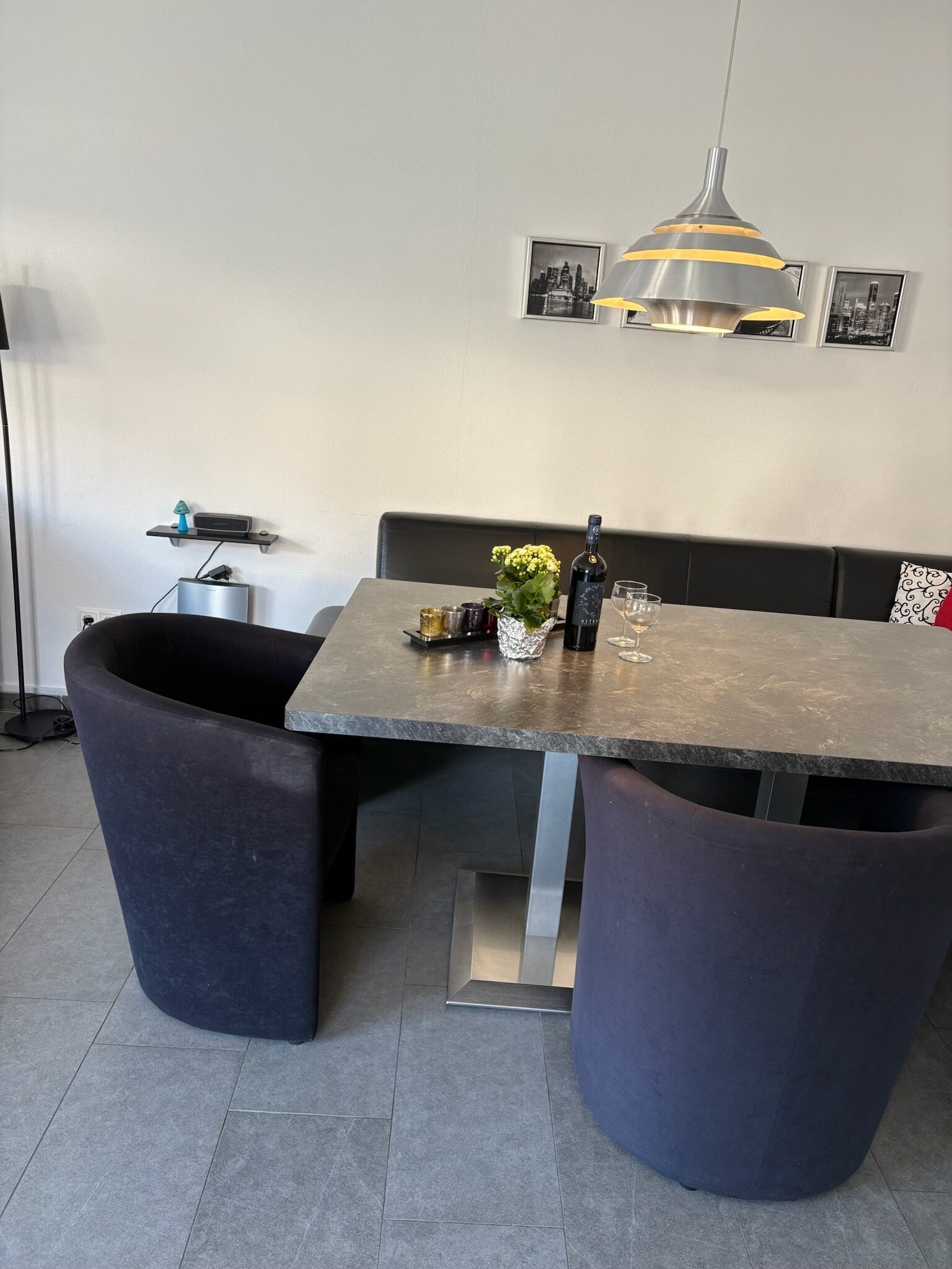 En moderne spiseplads med et gråt rektangulært bord med en sort polstret bænk og to mørke stole. På bordet står en flaske vin, to glas, en bakke og en lille potteplante. En pendel hænger over bordet.