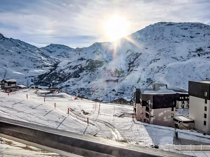 Solopgang over et snedækket bjerglandskab med skihytter og skilifte. Solen kaster et skarpt skær over snedækkede skråninger og tinder. Bygninger og skiløjper er synlige i forgrunden, omgivet af det store, fredfyldte vinterlandskab.