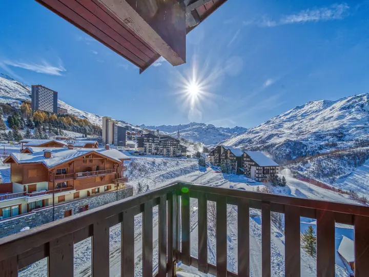 Udsigt fra en træbalkon med udsigt over et snedækket bjergresort. Solen skinner klart på en klar blå himmel og oplyser hytter og bygninger beliggende i det sneklædte landskab. Snedækkede tinder rejser sig i baggrunden.