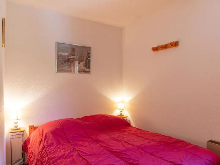 Et hyggeligt soveværelse med et rødt sengetæppe, to små natborde med lamper på hver side, et indrammet billede på væggen og en knagerække i træ monteret over sengen. Rummet har et blødt lys, der skaber en varm stemning.