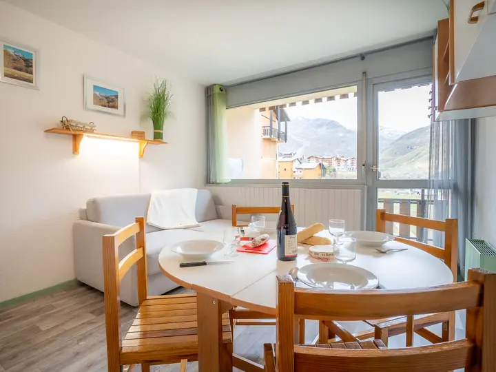 Lys, hyggelig spiseplads med et rundt bord til tre. Stort vindue tilbyder udsigt over bjergene. Hvid sofa, træstole og enkel indretning med indrammede billeder og en lille plante fremhæver den indbydende atmosfære.