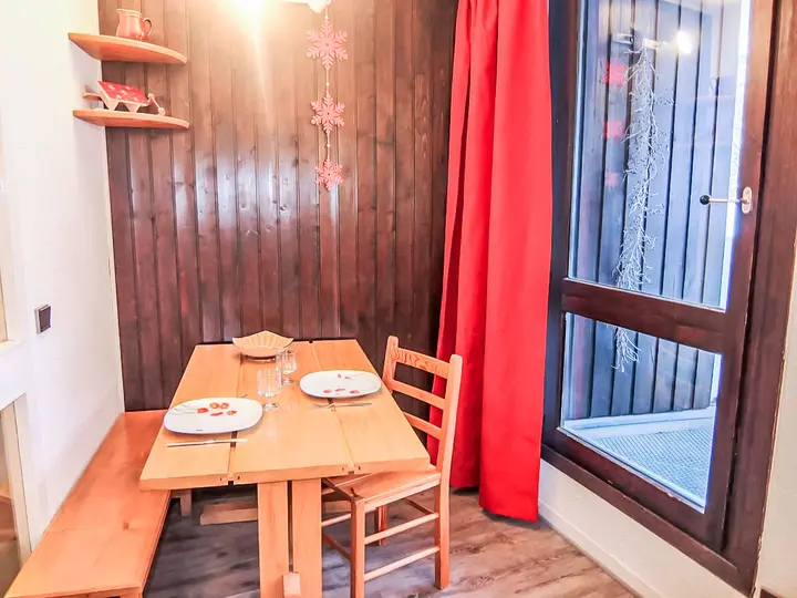 En lille hyggelig spiseplads med træbord og bænke. To tallerkener og nogle glas stilles på bordet. Væggen er af mørkt træ, dekoreret med røde accenter, og et stort vindue er prydet med et rødt gardin.