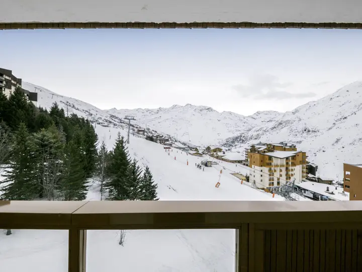 Udsigt fra en balkon med udsigt over et snedækket skisportssted. Forgrunden viser et trærækværk, mens skiløbere og bygninger er synlige på baggrund af snedækkede skråninger og fyrretræer. Himlen er klar med en antydning af skyer.