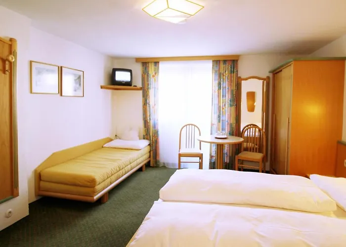Et lyst hotelværelse med en dobbeltseng, en enkelt sovesofa, et lille rundt bord med to stole ved et vindue med farverige gardiner, et klædeskab i træ og et lille tv på en væghængt hylde.
