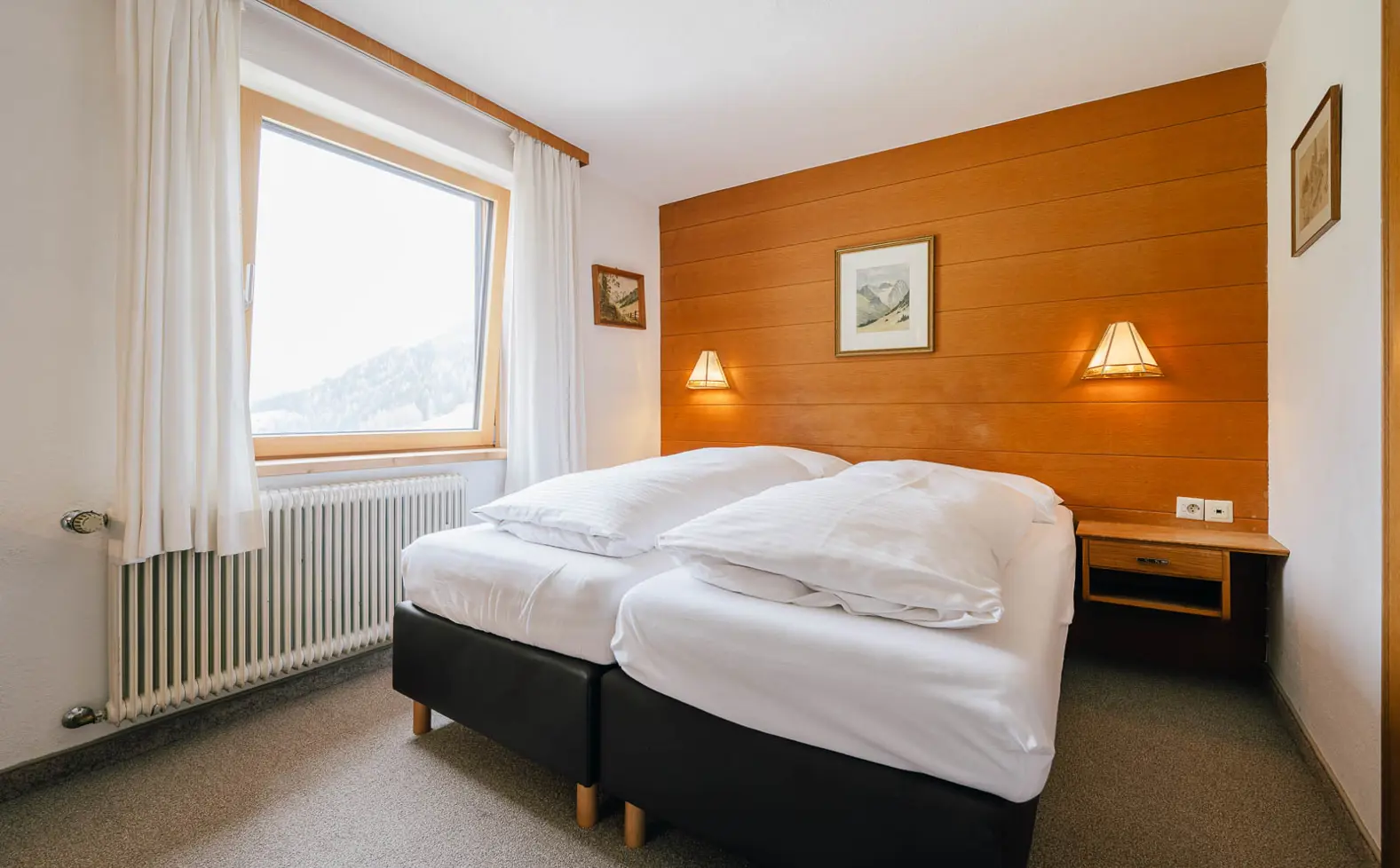 Et hyggeligt hotelværelse med to senge skubbet sammen, hvidt sengetøj, en træ-accentvæg, to væglamper, et vindue med hvide gardiner og indrammede billeder på væggene.