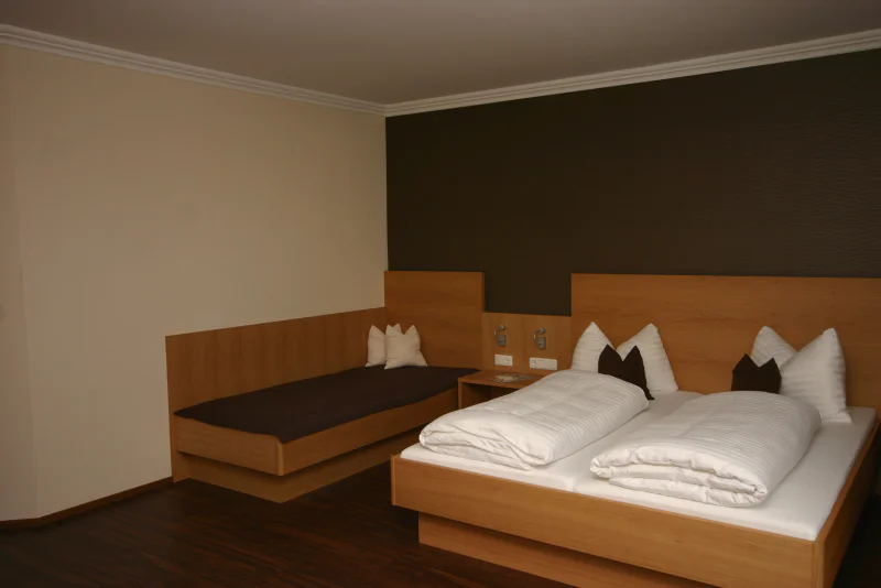 Et minimalistisk hotelværelse med en dobbeltseng og en enkeltseng, begge med trærammer, hvidt sengetøj og brune accentpuder, sat mod neutrale vægge og mørkt trægulv.