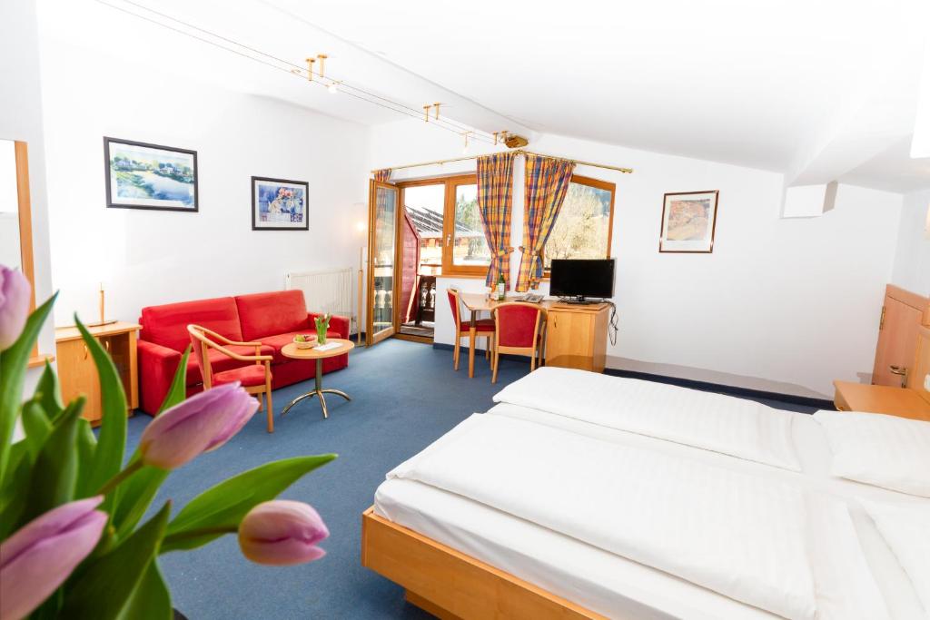 Lyst hotelværelse med dobbeltseng, rød sofa, lille bord med stole, skrivebord med tv, stort vindue med gardiner, billeder på hvide vægge, blåt tæppe og en vase med lyserøde tulipaner i forgrunden.