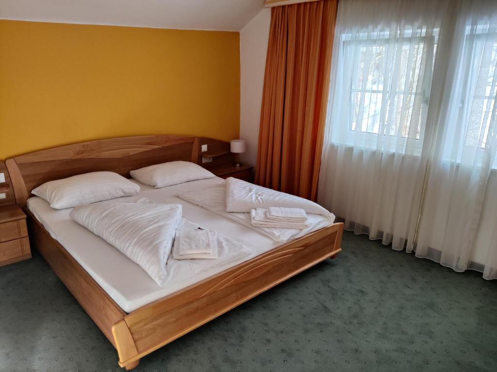Et pænt indrettet hotelværelse har en dobbeltseng i træ med hvidt sengetøj og foldede håndklæder på toppen. Væggene er malet halvt gule og halvt hvide. Et stort vindue med gennemsigtige gardiner og orange gardiner lukker naturligt lys ind. Grønt tæppe dækker gulvet.