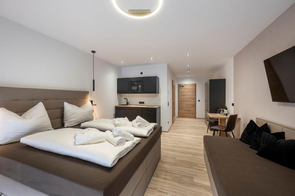 Moderne hotelværelse med stor seng, foldede håndklæder, sofa, væghængt tv, lille spisebord med stole og tekøkken. Rummet har trægulve og neutrale toner, med en dør yderst.