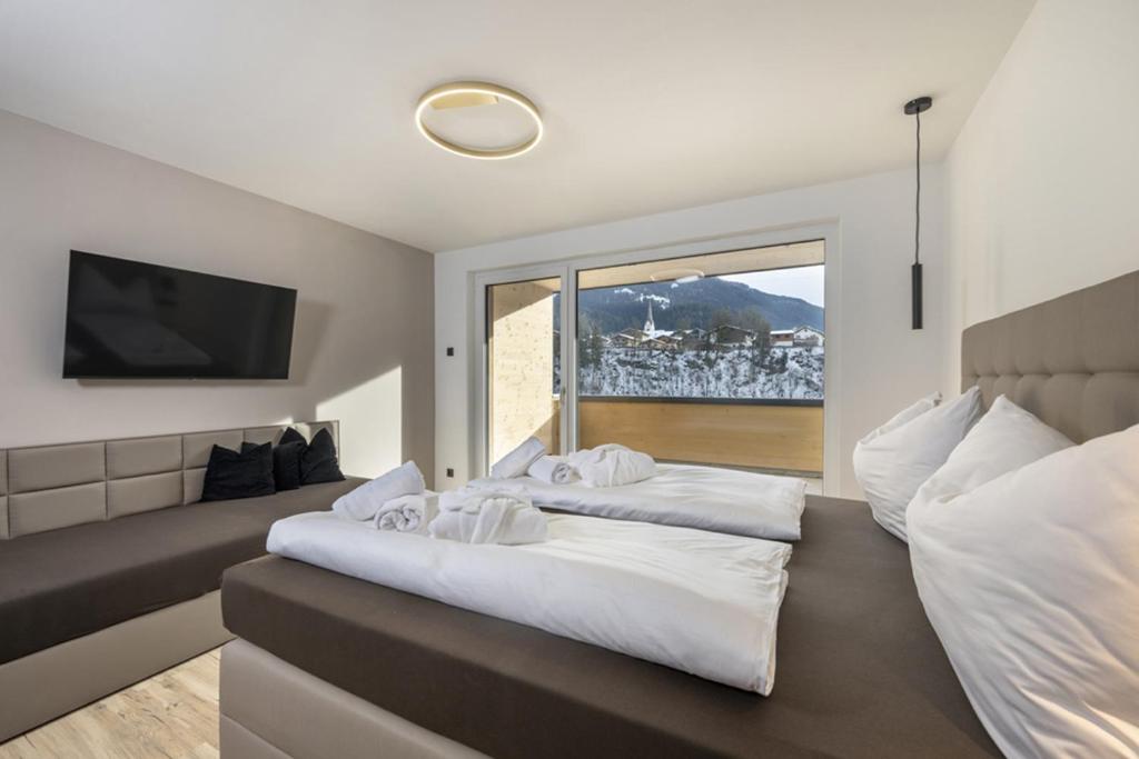 Moderne hotelværelse med pænt rede senge, et væghængt tv, en sofa med sorte puder og store vinduer, der viser et sneklædt bjerglandskab udenfor. Værelset er lyst og minimalistisk indrettet.