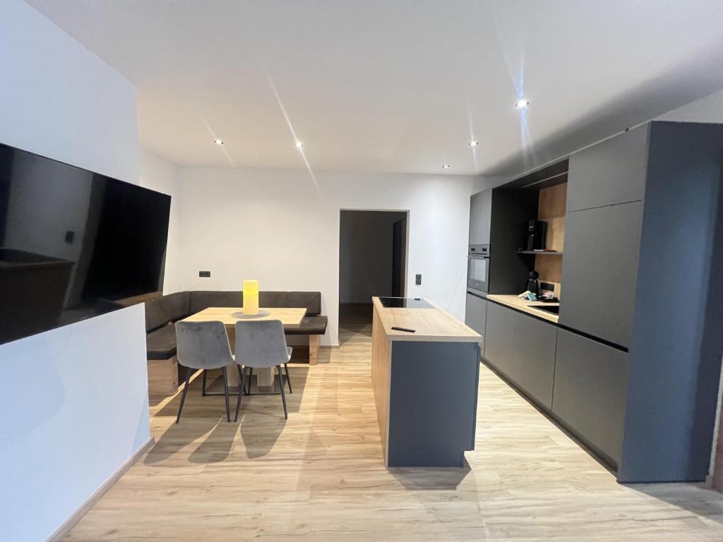 Moderne åbent køkken og spiseplads med grå skabe, en træø, en hjørnesofa, et spisebord med fire stole, lyse trægulve og minimalistisk indretning. Kraftig belysning fremhæver det rene design.