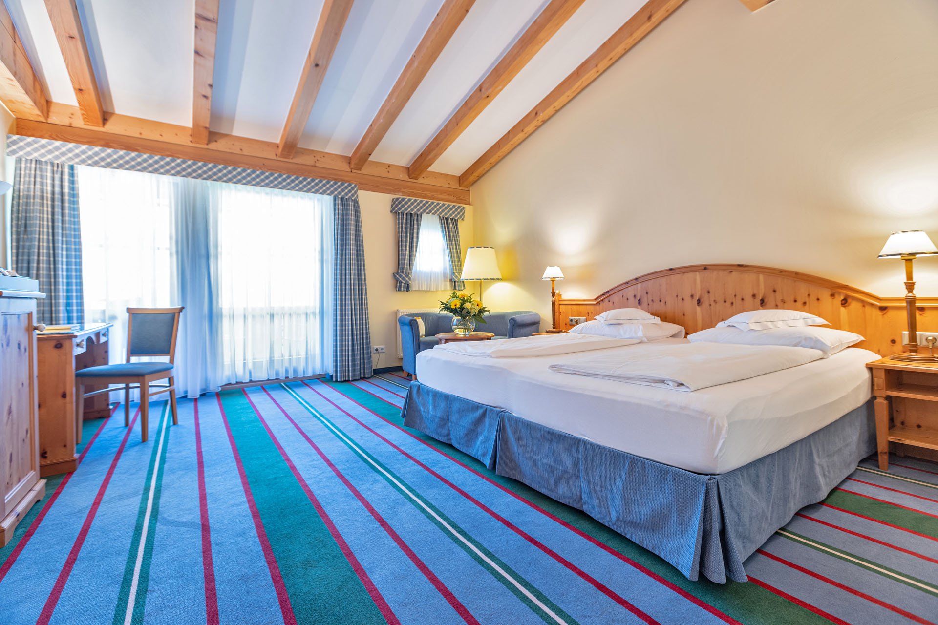 Lyst hotelværelse med træbjælker, stor dobbeltseng og blåstribet tæppe. To sengelamper, et skrivebord med en stol og to lænestole tæt ved et vindue med plaidgardiner fuldender de hyggelige rammer.
