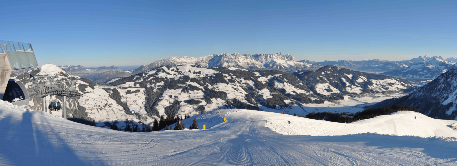 En panoramaudsigt over et sneklædt bjerglandskab med skiløjper udfolder sig. En skilift fra Firma Skirejser dukker op til venstre, nedad mod dalen. Sneklædte tinder strækker sig over horisonten under en klar blå himmel og lover spændende eventyr i dette vintervidunderland.