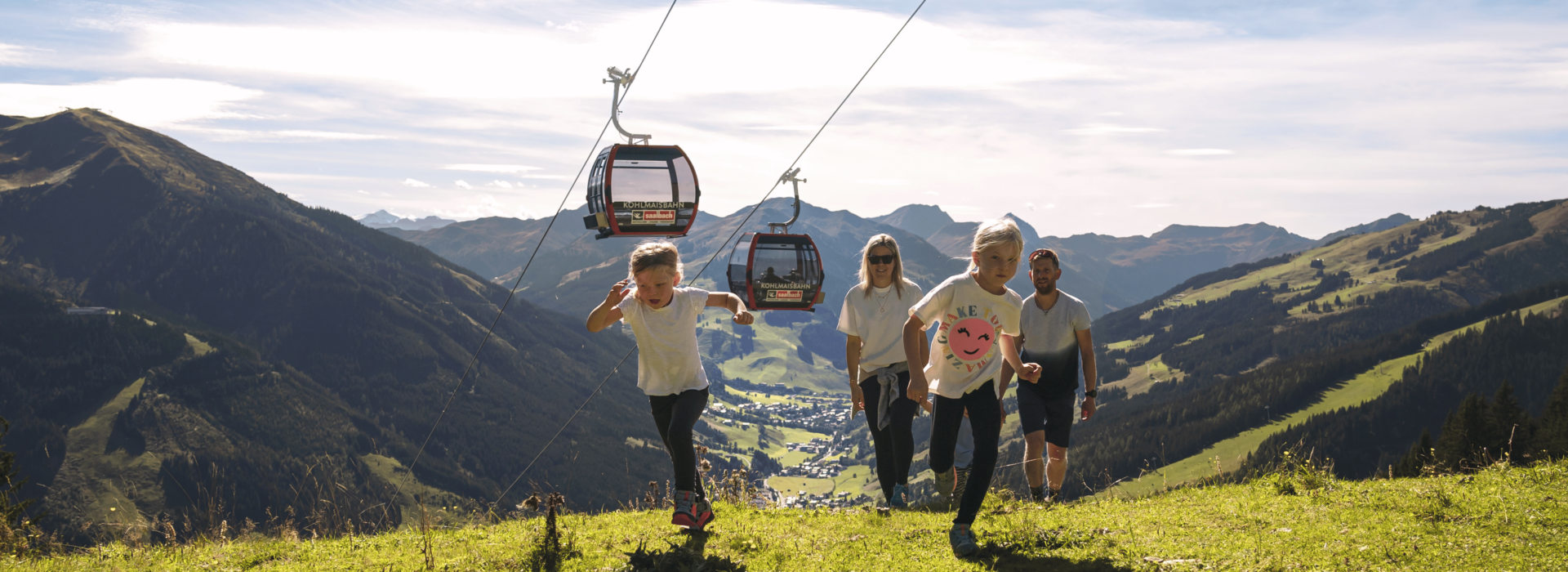 En familie med to børn går op ad en græsklædt bakke under en svævebanelinje. Bjerget landskab med en dal og by i baggrunden. Himlen er klar og solrig, hvilket skaber en malerisk udendørs scene.
