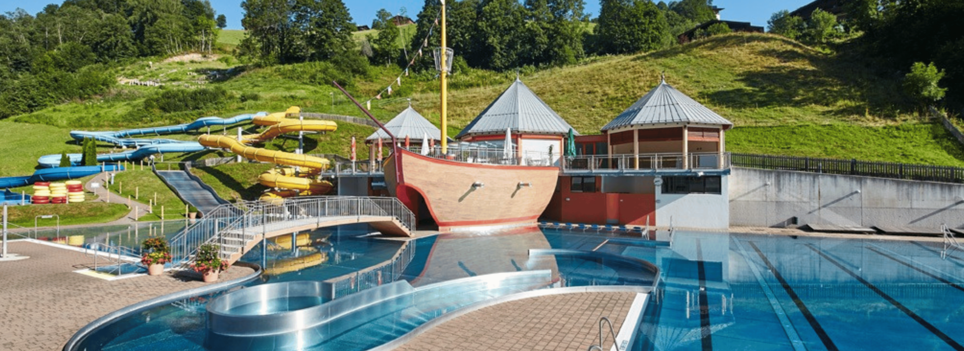 Udendørs vandland med en stor pool med blåt vand, en bro og en central struktur med piratskibstema. Forskellige farverige vandrutsjebaner snor sig ned ad en græsklædt bjergskråning i baggrunden. Træer og en klar himmel er synlige.